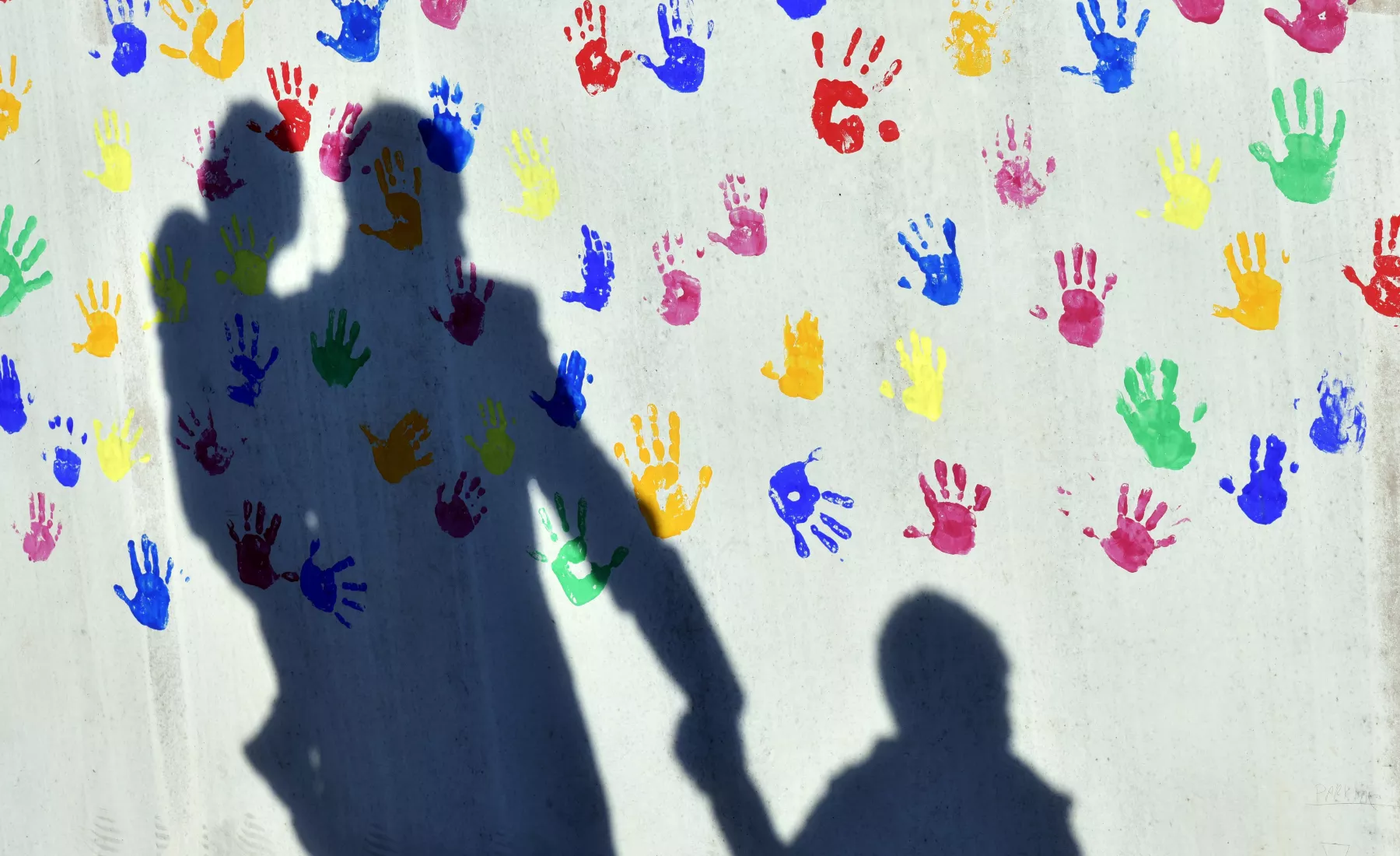 Ein Mann mit einem Kind auf dem Arm und einem an der Hand wirft einen Schatten auf eine mit bunten Handabdrücken bemalte Wand einer Kindertagesstätte