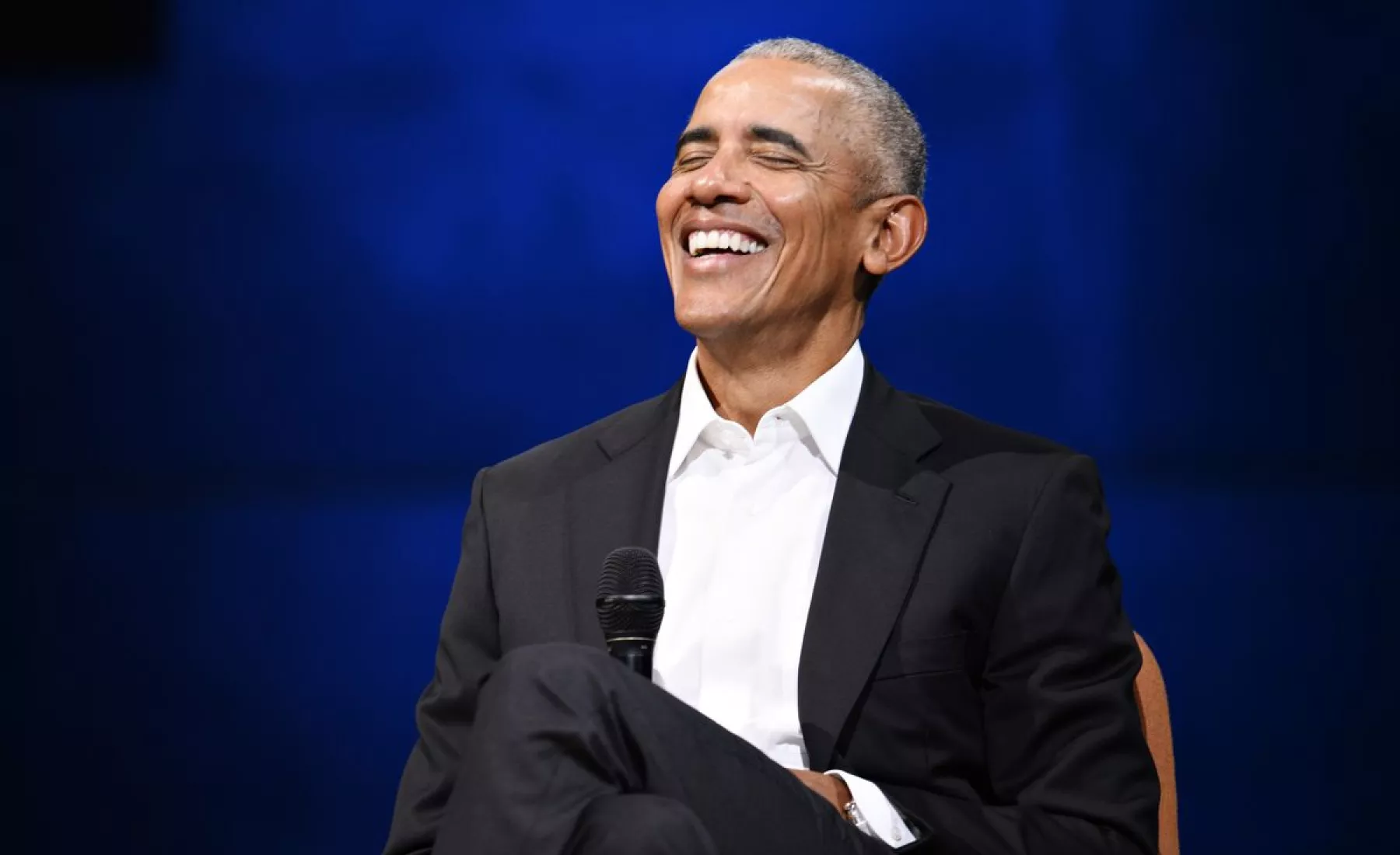 Barack Obama sitzt lachend auf einer Bühne mit einem Mikrofon in der Hand