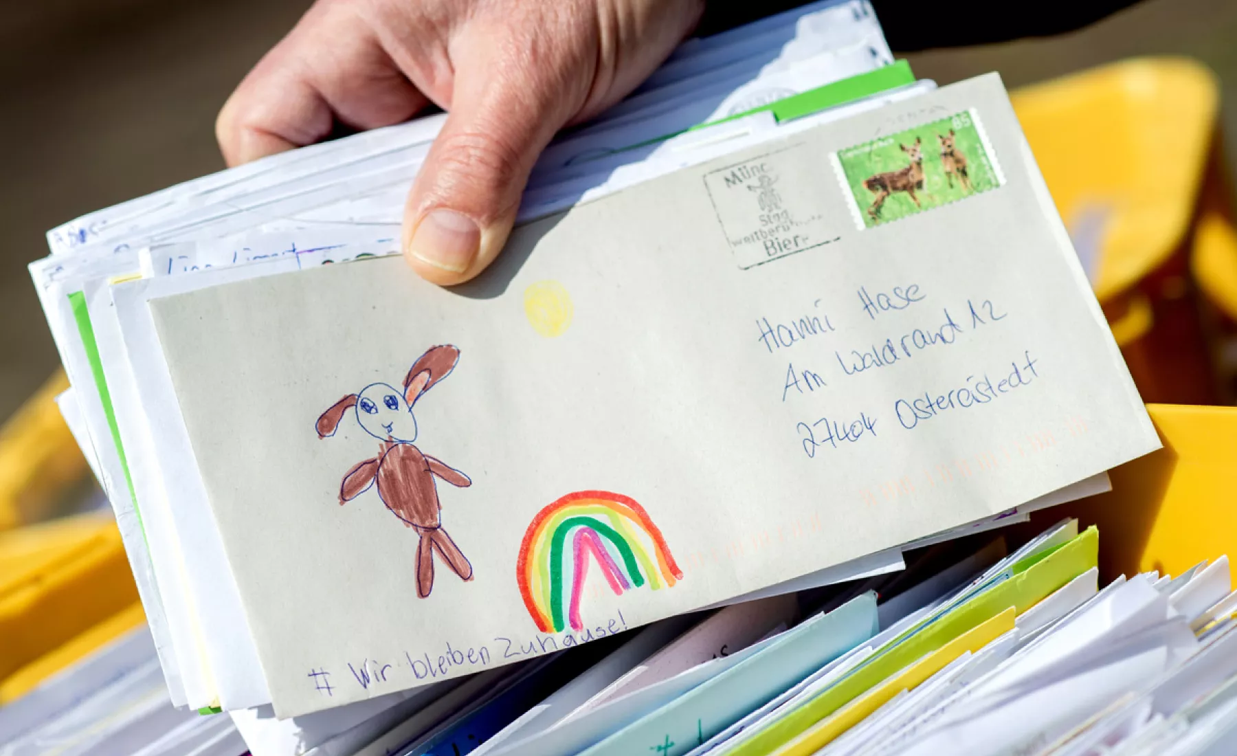 Ein Mitarbeiter des Osterpostamts hält vor dem Gemeindehaus von Ostereistadt einen Brief in den Händen, der einen gemalten Regenbogen und die Aufschrift «Wir bleiben Zuhause!» zeigt.