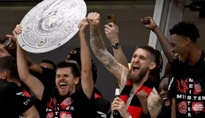 ie Leverkusener Mannschaft jubelt mit einer stilisierten Meisterschale von der Tribüne ins Stadion, den Fans zu, die nach dem Sieg das Spielfeld gestürmt haben.