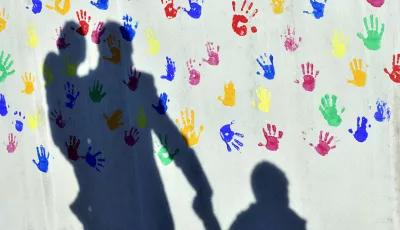 Ein Mann mit einem Kind auf dem Arm und einem an der Hand wirft einen Schatten auf eine mit bunten Handabdrücken bemalte Wand einer Kindertagesstätte