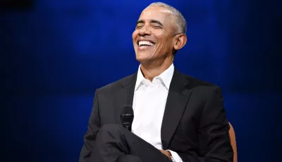 Barack Obama sitzt lachend auf einer Bühne mit einem Mikrofon in der Hand