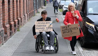 Eine Frau und ein Mann im Rollstuhl gehen mit Plakaten „Unschuldige behinderte Menschen töten. Warum?“ und „Unser aufrichtiges Beileid“ zum Eingang der Einrichtung des diakonischen Anbieters Oberlinhaus.